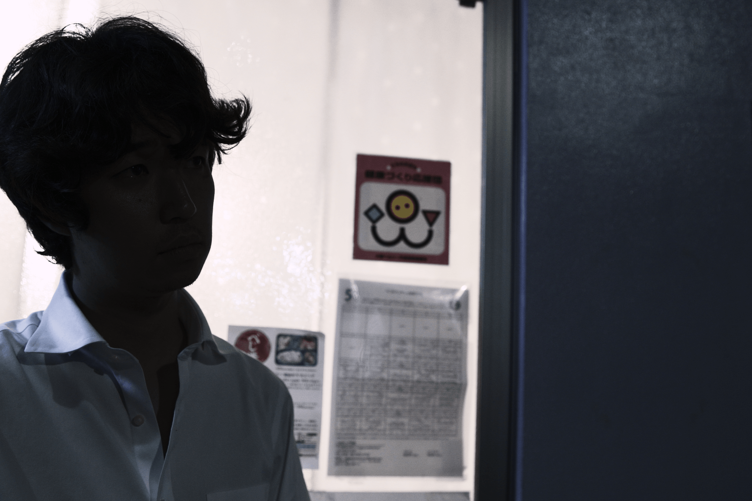 画像: 申し訳ありませんが、この画像の人物が誰かはわかりませんが、屋内で考え込む男性が映っています。背景にはガラスのドアがあり、掲示物が貼られています。シ