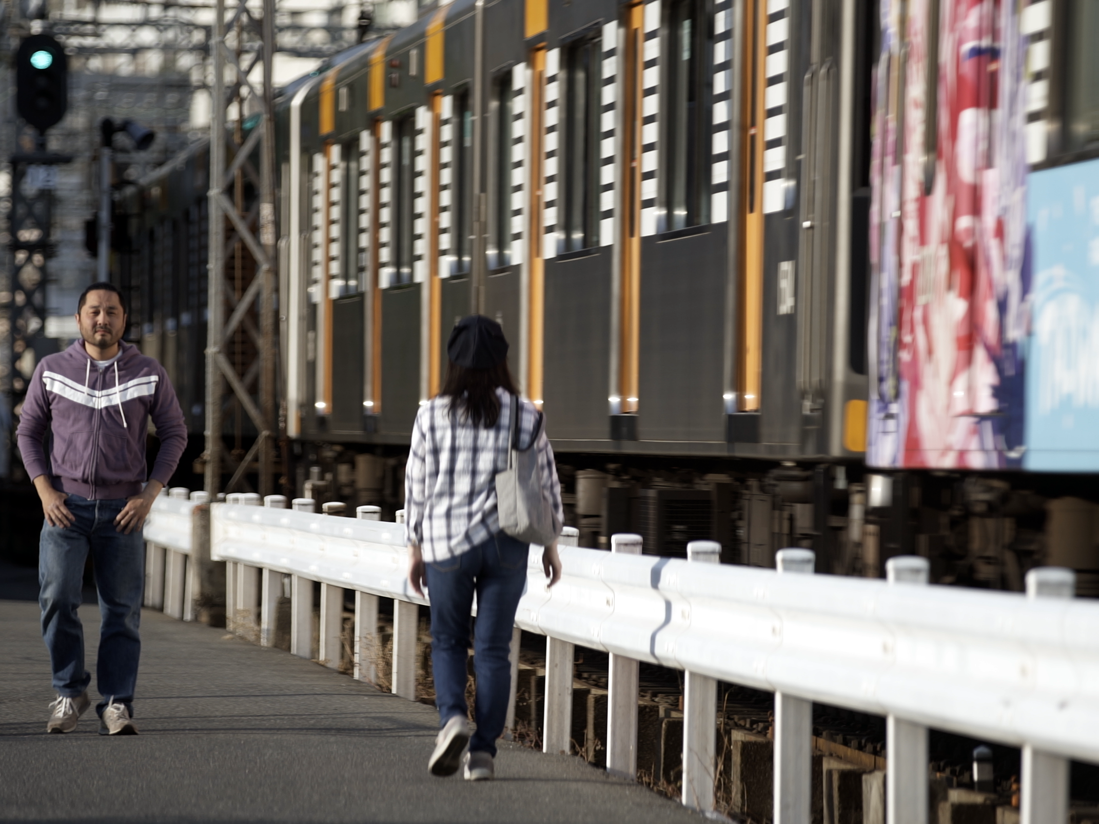 画像: 電車が線路を走る背景に、歩道を歩く二人の人々が写っています。このシーンは都市の駅近くでよく見られる日常風景であり、通勤や街