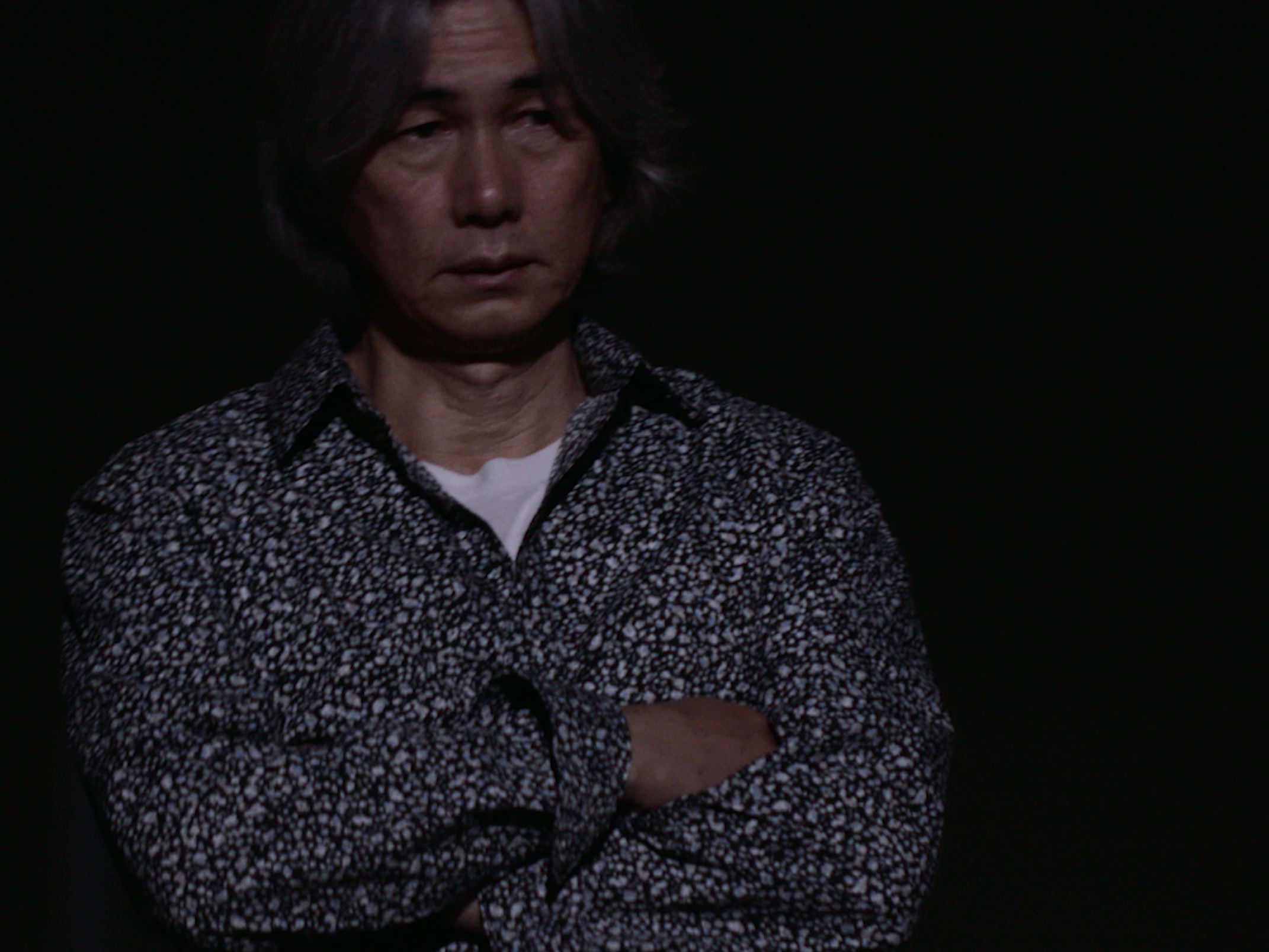 画像: 申し訳ありませんが、画像内の人物が誰かを特定することはできません。この画像では、暗い背景にシックな柄のシャツを着た男性が腕を組んで立っています。照明