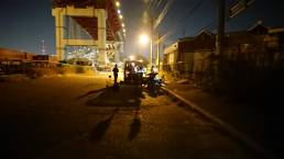 画像: 夜の都市で、工業地帯の高架下に集まって作業している人々がいます。薄暗い中、車のライトで照らされた様子が印象的です。キーワード: 夜