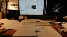 画像: オフィスデスクの画像には、2台のコンピュータモニターと、印刷された資料が散らばったデスクがあります。デジタル時計やスピーカーも見えます。作業