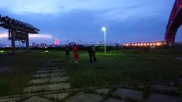 画像: 夜の公園で数人が談笑しているシーンです。背景には観覧車と橋がライトアップされ、大阪湾沿いの風景が広がっています。大坂の夜景、観覧車、