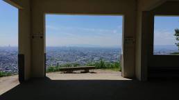 画像: コンクリートの建物の窓枠から大阪の街並みを一望する風景。青空が広がり、地平線に都市のスカイラインが見える。場所は高台