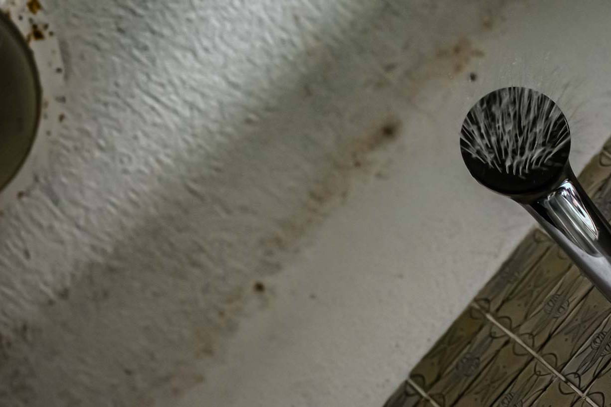 画像: バスルームの壁にあるシャワーヘッドが水を出しているシーンです。背景には古いタイルと白い壁が見えます。「シャワー」、「バスルーム」、「水流」、「