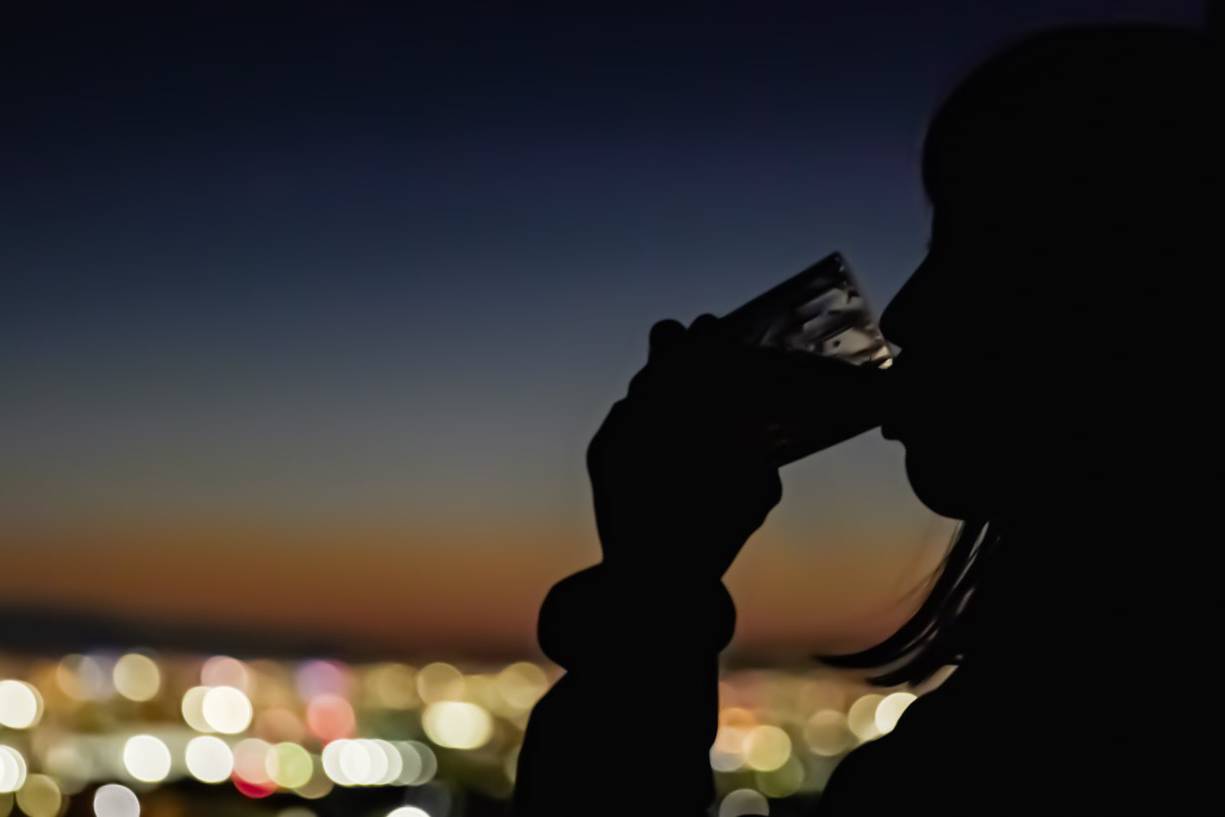 画像: 夕暮れ時の都市の夜景を背景に、飲み物を手にする人物のシルエット。美しいボケ効果が都市の光を際立たせ、ロマンチックな雰