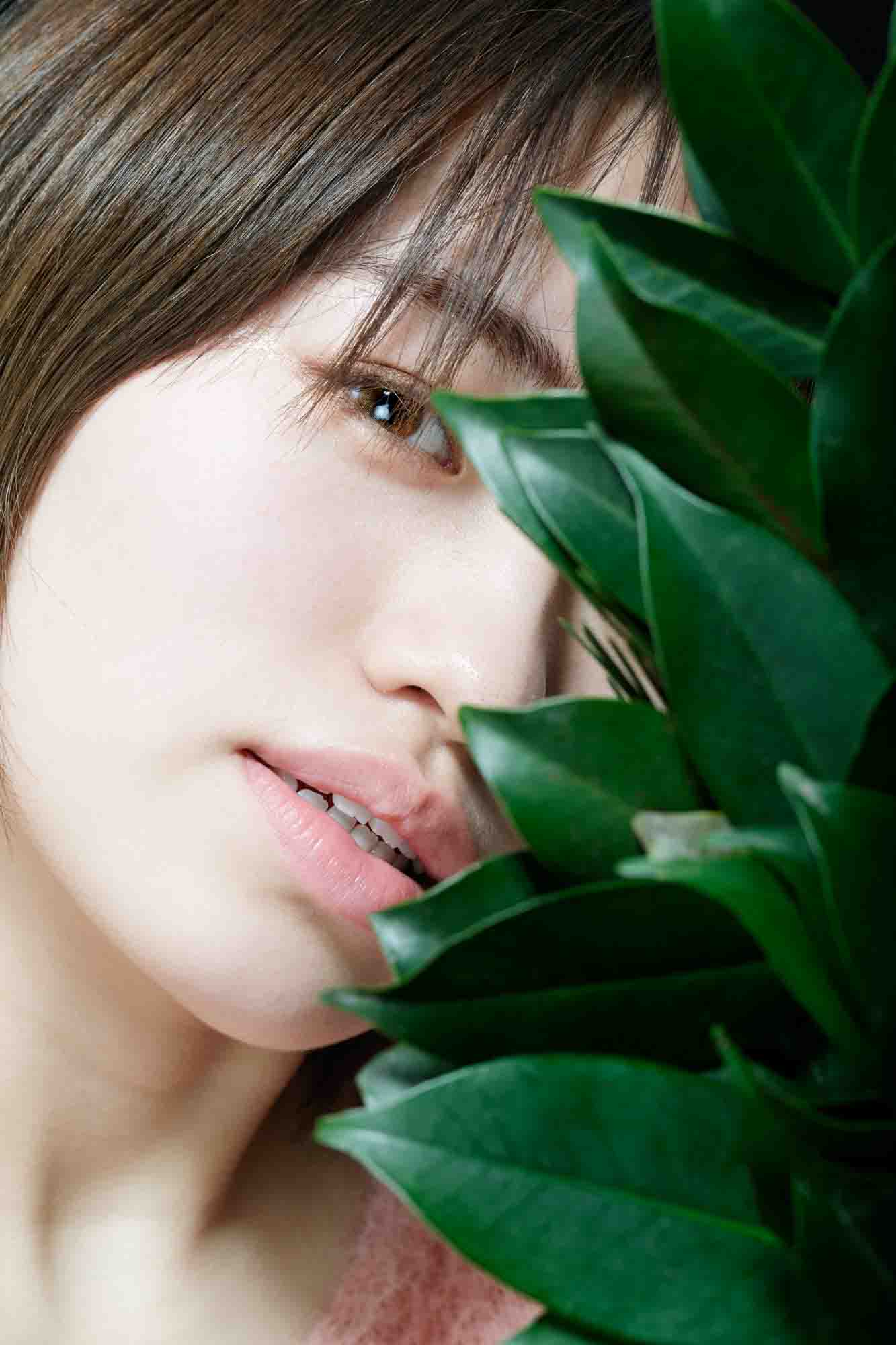 画像: 若い女性が植物の葉の後ろから覗いているクローズアップ写真です。背景はシンプルで、ナチュラルな美しさを強調しています。関連するキーワードは、ポ