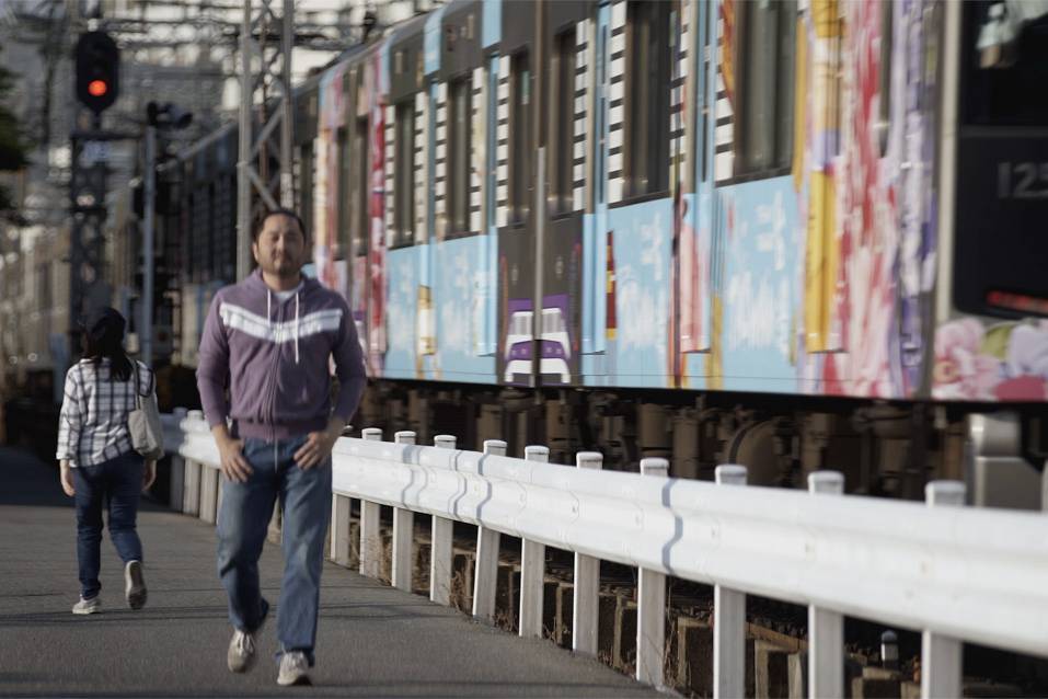 画像: 鉄道沿いを歩く人々とカラフルな外観の電車。東京都内で撮影された都市の風景。鉄道、駅、都市生活、歩行者のキーワードが考えら