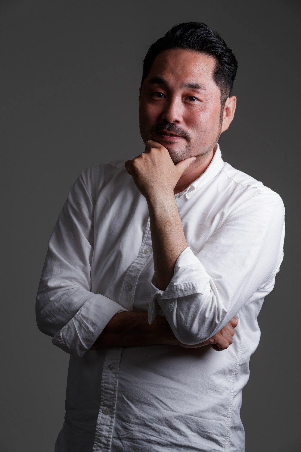 画像: 白いシャツを着た男性が腕を組み、手を顎に当てて考え事をしているポートレートです。落ち着いた雰囲気が漂うスタジオ撮影