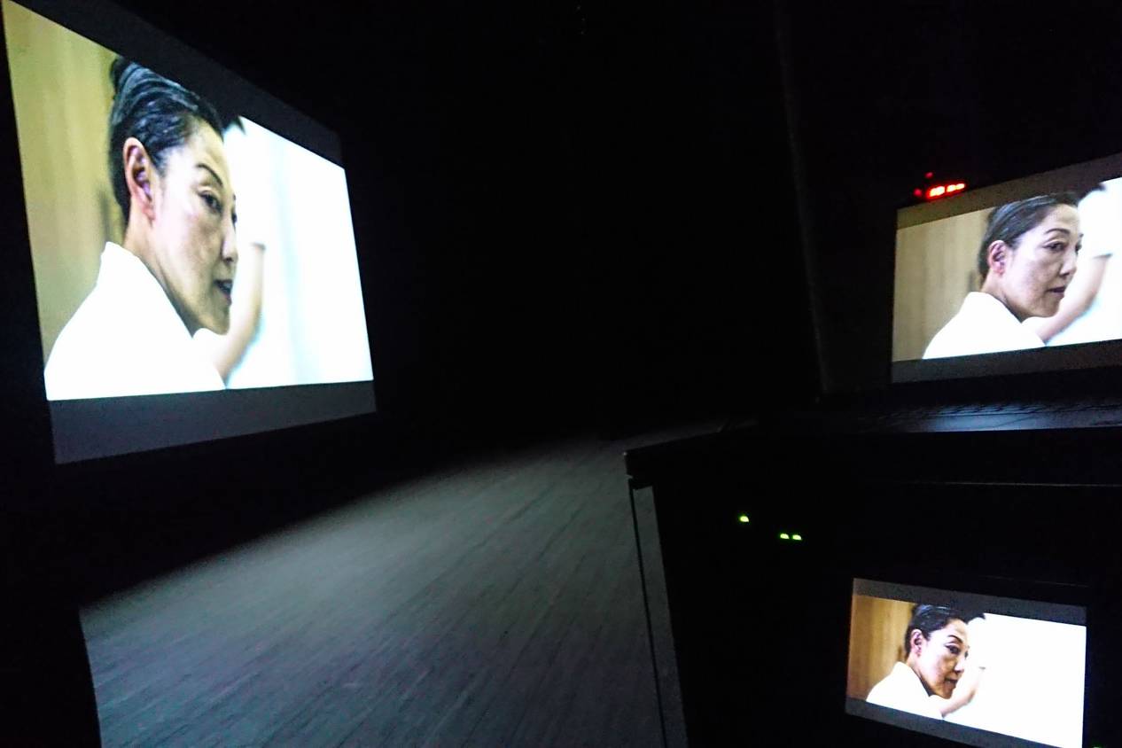 画像: 劇場内で映画が上映されている様子。スクリーンと複数のモニターに同じシーンが映し出され、暗い空間が強調されています。キーワード: 映画