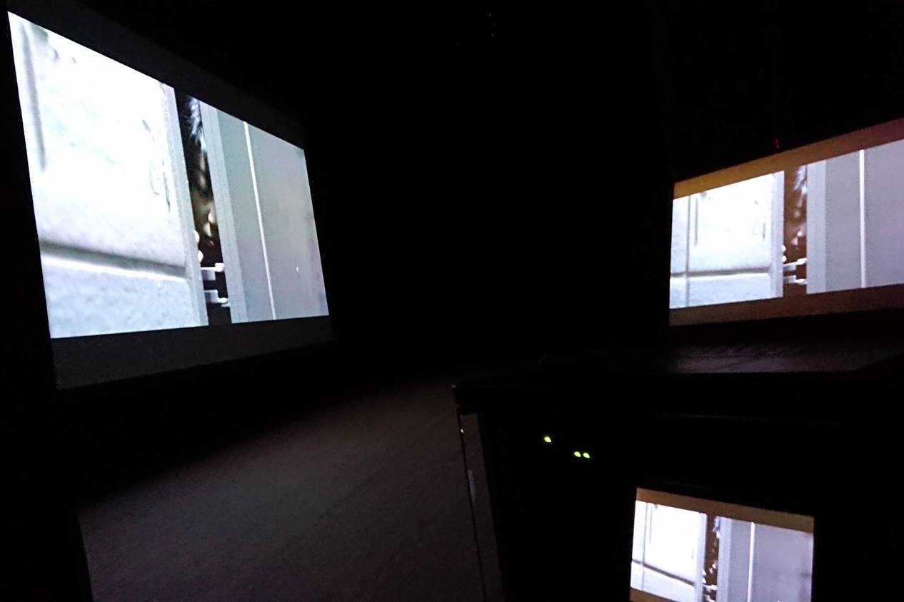 画像: 暗い部屋でプロジェクターとスクリーンに映し出された映像が映るシーン。壁に大きく投影された画像と、近くのモニターに同じ映像が表示