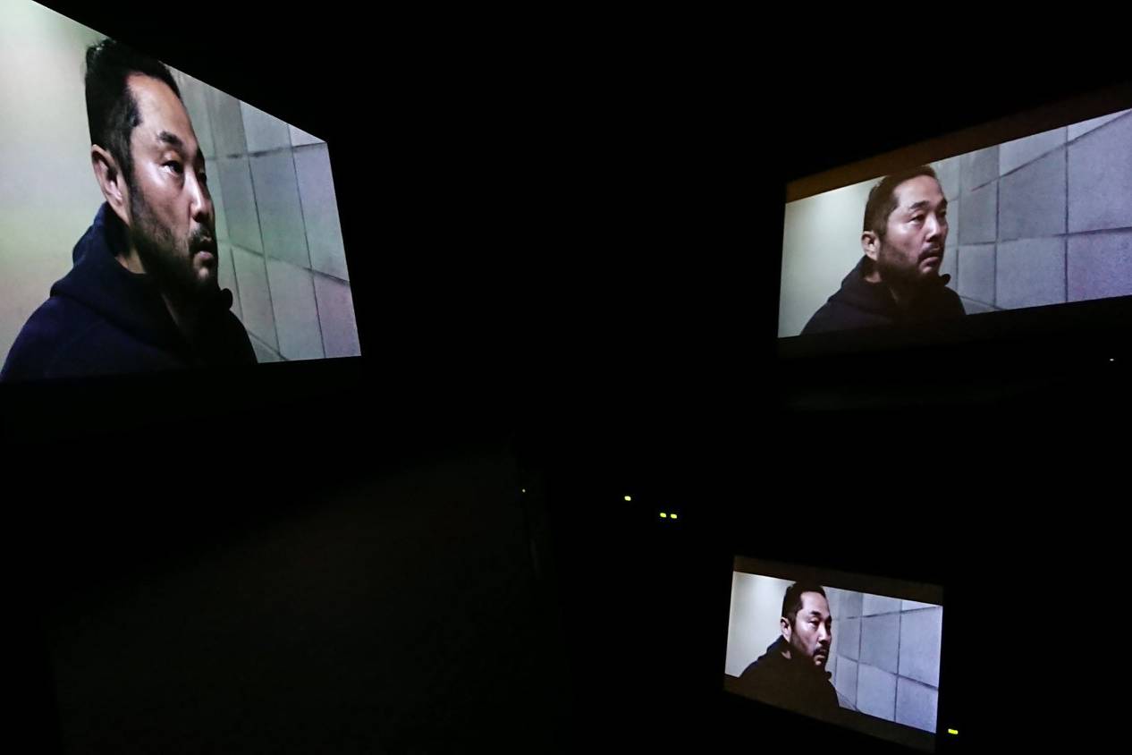 画像: スクリーンに複数映し出された映画のシーンで、男性が壁際に立っています。ダークな雰囲気が支配するこの映像は、サスペンス映画やスリラー、