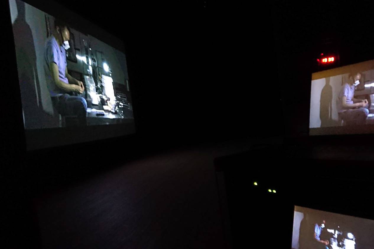画像: この画像は、暗闇の中で映画がプロジェクターから上映されているシーンを示しています。スクリーンには作業中の人物が映し出されています。映画館、映写機、上映
