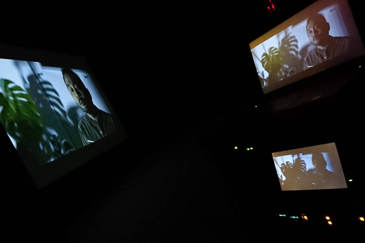 画像: 画像には、暗い部屋で複数のスクリーンに映し出された男性の映像が写っています。スクリーンには観葉植物が背景として映っています。映画鑑賞や家庭用プロジェ