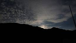 画像: 山のシルエットと雲の動きが特徴的な夕暮れの空の風景です。逆光の中で空が美しく描かれており、静寂な自然が広がります